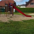Ein Spielplatz mit Klettergerüst, Rutsche und Wippe, umgeben von Rasen und modernen Wohnhäusern.