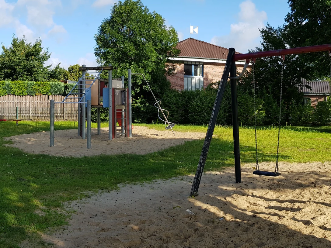 Spielplatz mit Rasenfläche, Klettergerüst und Schaukeln auf Sandboden, umgeben von grünen Bäumen.