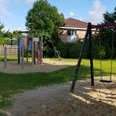 Spielplatz mit Rasenfläche, Klettergerüst und Schaukeln auf Sandboden, umgeben von grünen Bäumen.
