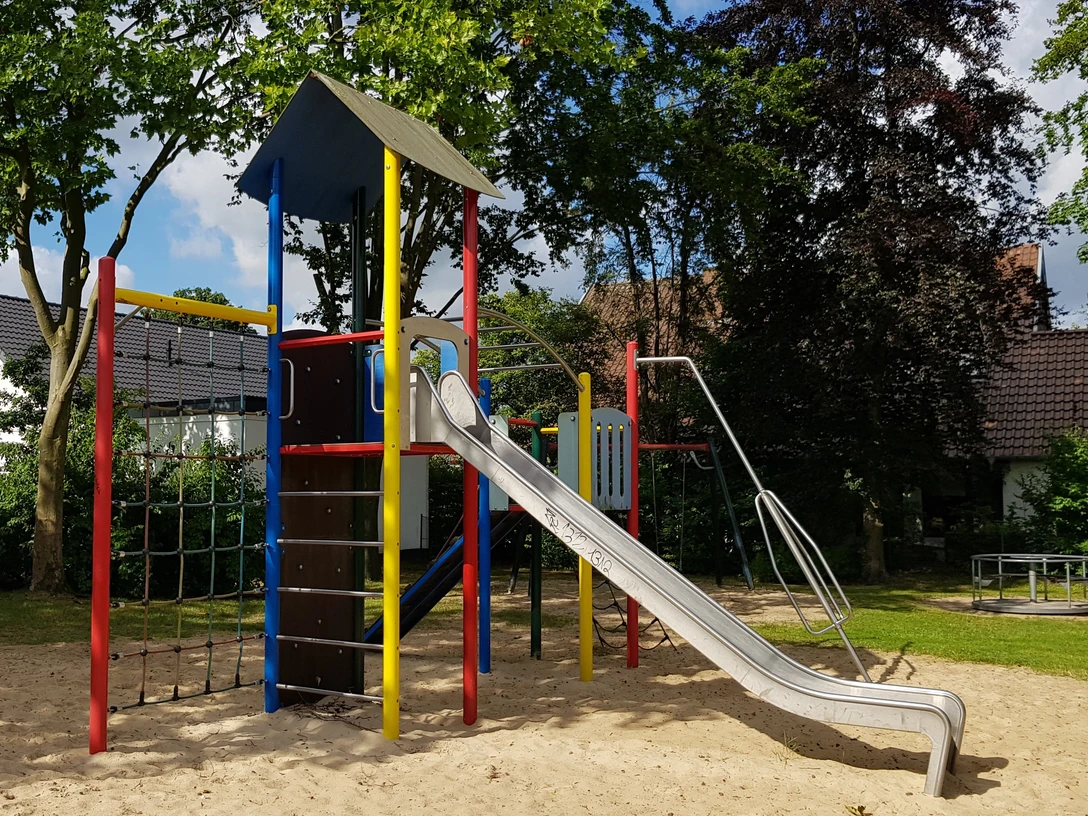 Spielplatz Pirolweg Moderner Spielplatz mit buntem Klettergerüst und Rutsche, umgeben von grünen Bäumen und Rasenflächen.