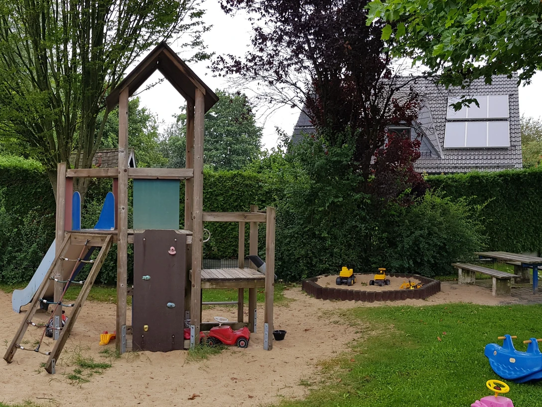 Spielplatz mit Klettergerüst, Rutsche und Sandkasten; umgeben von grünem Gras und Bäumen, Haus im Hintergrund.