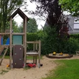 Spielplatz Masurenstraße Spielplatz mit Klettergerüst, Rutsche und Sandkasten; umgeben von grünem Gras und Bäumen, Haus im Hintergrund.