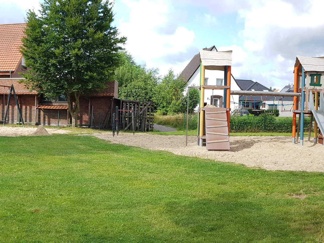 Spielplatz Jostweg Spielplatz mit grüner Wiese, zwei Rutschen, Schaukeln und umgeben von Häusern bei bewölktem Himmel.