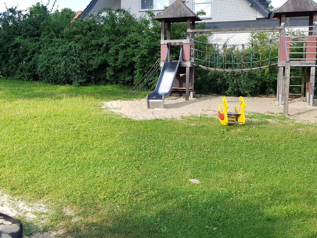 Spielplatz mit Rutsche, Klettergerüst und Wippspielzeug auf sandigem Boden, umgeben von Rasen.
