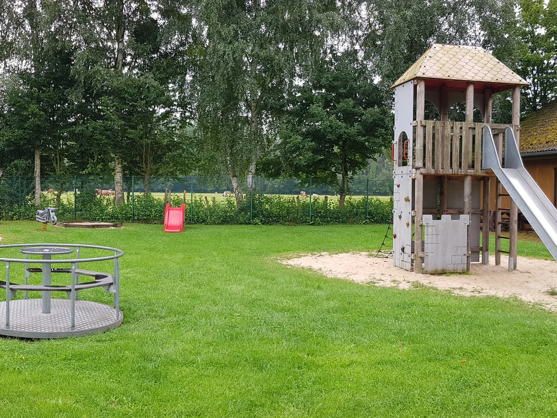Spielplatz Kieselweg Ein grüner Spielplatz mit Kletterturm, Rutsche, Karussell und Schaukeln, umgeben von Bäumen.