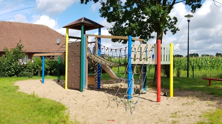 Ein Spielplatz mit Klettergerüst, Rutsche und Kletternetzen auf sandigem Untergrund, umgeben von Natur.