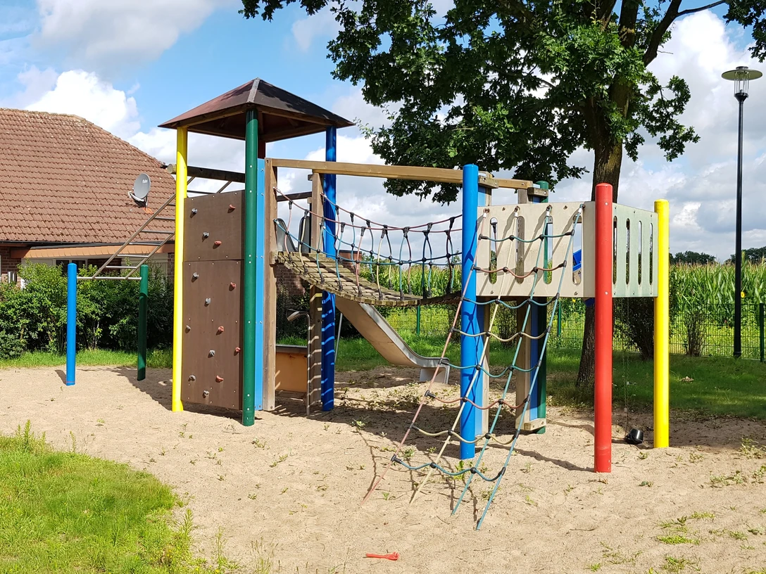 Ein Spielplatz mit Klettergerüst, Rutsche und Kletternetzen auf sandigem Untergrund, umgeben von Natur.