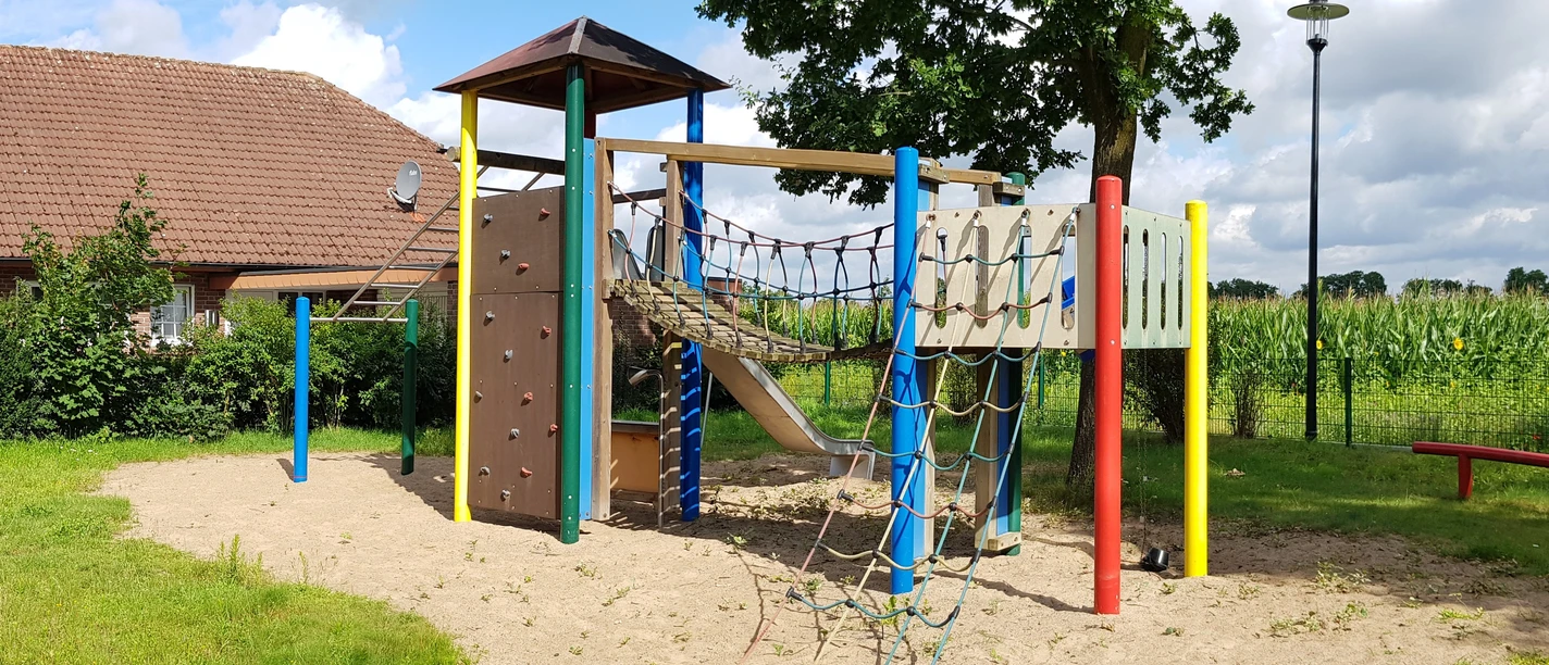 Ein Spielplatz mit Klettergerüst, Rutsche und Kletternetzen auf sandigem Untergrund, umgeben von Natur.