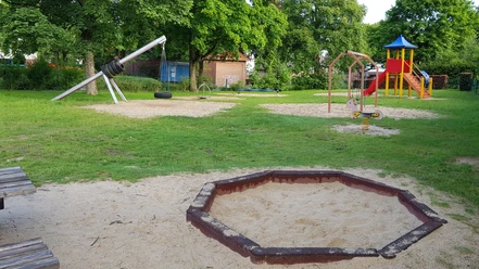 Spielplatz Lessingstraße Ein grüner Spielplatz mit Klettergerüst, Wippe und Schaukeln, umgeben von Bäumen und Sandflächen.