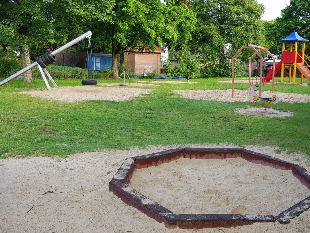 Spielplatz Lessingstraße Ein grüner Spielplatz mit Klettergerüst, Wippe und Schaukeln, umgeben von Bäumen und Sandflächen.