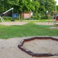 Spielplatz Lessingstraße Ein grüner Spielplatz mit Klettergerüst, Wippe und Schaukeln, umgeben von Bäumen und Sandflächen.