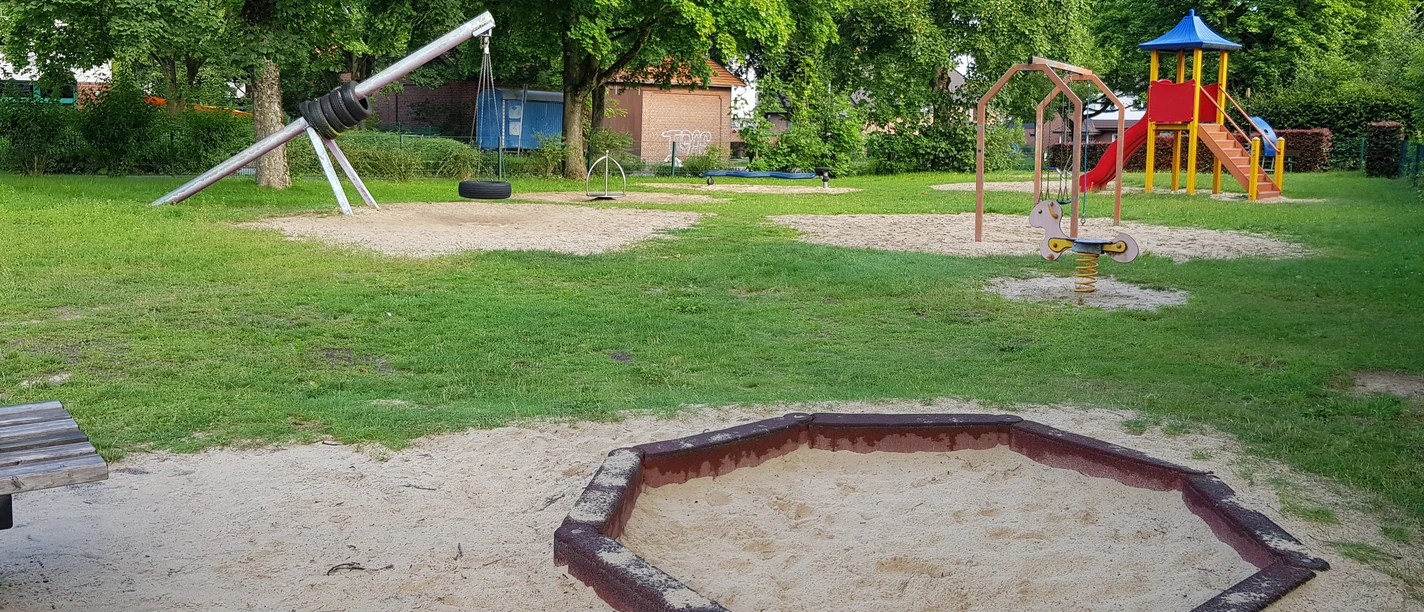 Spielplatz Lessingstraße Ein grüner Spielplatz mit Klettergerüst, Wippe und Schaukeln, umgeben von Bäumen und Sandflächen.