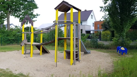 Spielplatz in Wohngegend mit Rutsche und Klettergerüst auf Sandfläche, umgeben von Bäumen und Häusern.