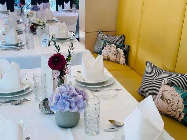Eleganter, festlich gedeckter Tisch in einem Restaurant mit weißen Tischdecken, Blumen und gelber Sitzbank.