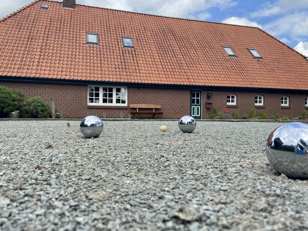 image00009.jpeg Ein rustikales Backsteingebäude mit rotem Ziegeldach im ländlichen Stil; im Vordergrund ein Boulespielfeld mit glänzenden Kugeln und einem kleinen Zielball.