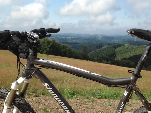 Waldsofa am Blockhaus Blick von einer Holzbalkenbank: Ein Mountainbike und weite, hügelige Landschaft unter blauem Himmel.