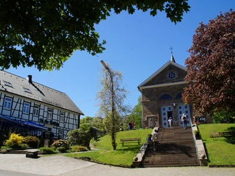 Kirche in Kürten-Delling Eine steinerne Kirche mit Treppe, daneben ein Fachwerkhaus, grüne Bäume und blauer Himmel.