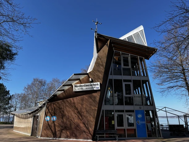 Moderne bistro met opvallende architectuur, glazen gevel en groot naambord, omringd door bomen.