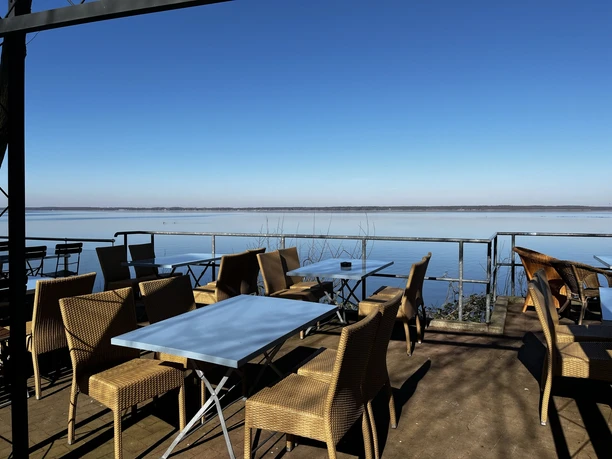 Bistro Meereslauschen Terrasse mit Rattanmöbeln am Wasser, klarer Himmel, ruhige Atmosphäre, Aussicht auf farblose See.