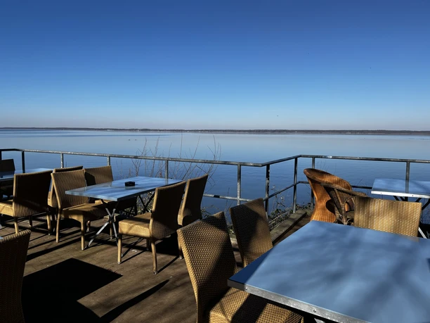 Ein Bistro mit Korbstühlen und blauen Tischen auf einer Terrasse, die an einem See gelegen ist.