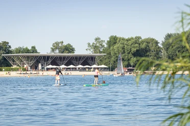 Menschen paddeln auf dem Allersee vor Strand, Segelboot und moderner Uferanlage im Allerpark.