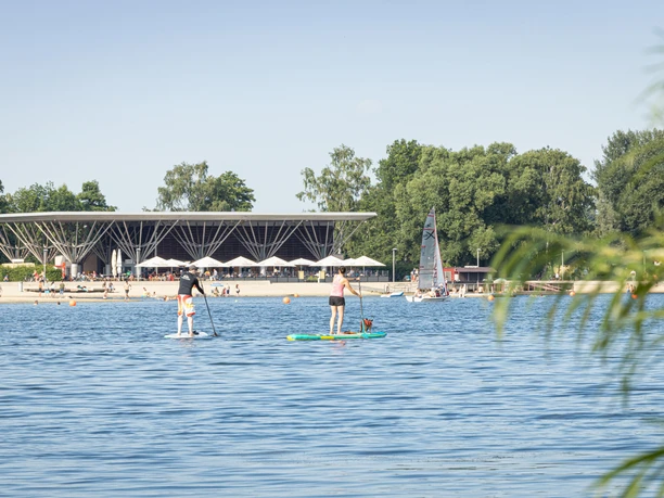 Menschen paddeln auf dem Allersee vor Strand, Segelboot und moderner Uferanlage im Allerpark.
