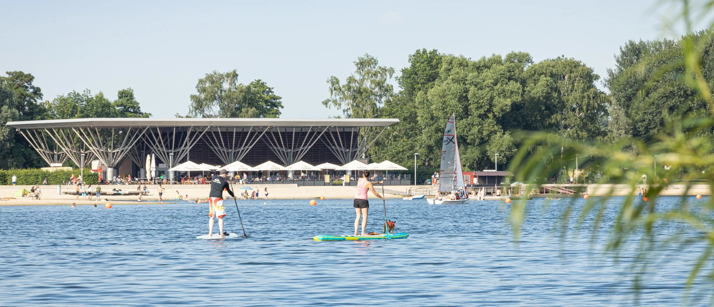 Allersee im Allerpark Menschen paddeln auf dem Allersee vor Strand, Segelboot und moderner Uferanlage im Allerpark.