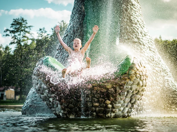 Trixi Ferienpark_Philipp Herfort (61-bearbeitet.jpg Ein Junge rutscht lachend eine Wasserfontäne hinunter, umgeben von spritzendem Wasser.