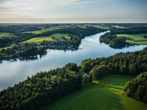Bevertalsperre Luftbild eines Sees in grüner Landschaft mit Wald, Feldern und einem Dorf am Ufer im Morgenlicht.