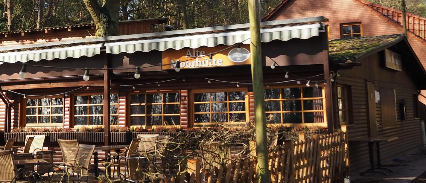 Alte Moorhütte Eine rustikale Holzhütte mit Veranda und Tischgruppe, umgeben von hohen Bäumen im Waldrestaurant.