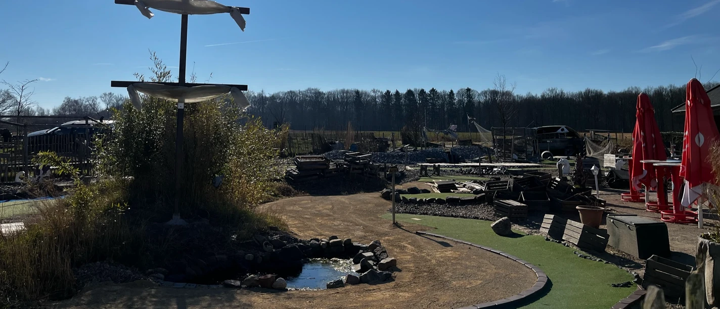 Blick auf das Gelände Activity Park Hagenburg Grünes Minigolfgelände im Activity Park Hagenburg mit Schirmen, Bänken und einem kleinen Teich.