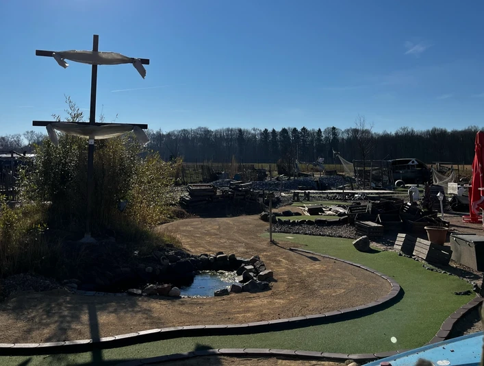 Blick auf das Gelände Activity Park Hagenburg Grünes Minigolfgelände im Activity Park Hagenburg mit Schirmen, Bänken und einem kleinen Teich.