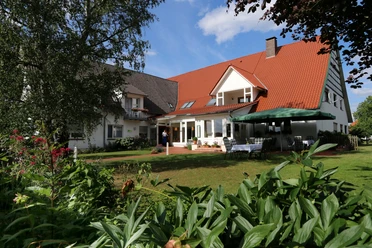 Landhaus Blankenstein Ein Landhaus mit roten Dachschindeln, umgeben von grünen Pflanzen und einem sonnigen, blauen Himmel.