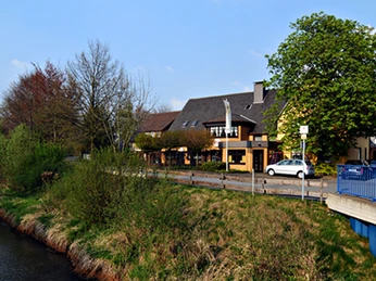 Gasthof Bökamp_Außenansicht Gasthaus am Fluss mit grünen Bäumen und blauer Brücke; ein idyllischer Frühlingstag.