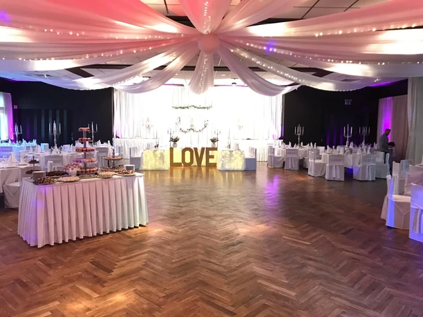 Gasthof Bökamp_Festsaal Festlich dekorierter Hochzeitssaal mit holzgemasertem Boden, weißen Tischen und großer "LOVE"-Leuchtschrift.