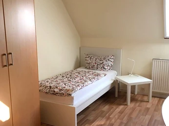 Ein gemütliches Einzelzimmer mit einem Bett, Holzboden, Schrank und einem kleinen weißen Nachttisch.
