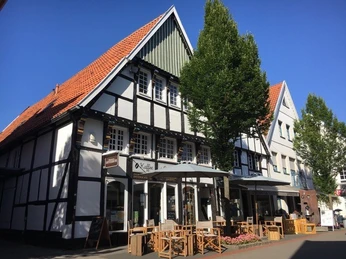 Kaffeekontor_1.jpg Fachwerkhaus mit Café, Holzmöbeln und Markisen in sonniger Einkaufsstraße; blauer Himmel, grüner Baum.