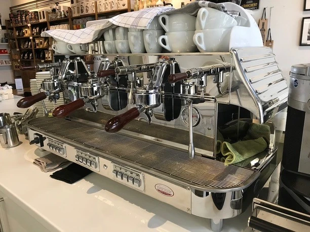 Eine silberne Espressomaschine mit mehreren Siebträgern steht auf einer weißen Theke in einem Café.