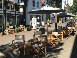 Kaffeekontor_4.jpg Ein Café mit verstellbaren Holzstühlen und Tischen unter großen Sonnenschirmen in einer Fußgängerzone.