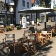 Kaffeekontor_4.jpg Ein Café mit verstellbaren Holzstühlen und Tischen unter großen Sonnenschirmen in einer Fußgängerzone.