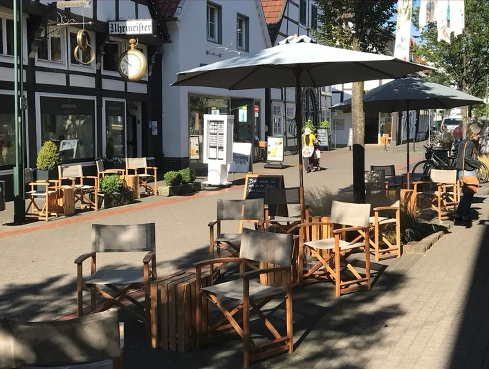 Kaffeekontor_4.jpg Ein Café mit verstellbaren Holzstühlen und Tischen unter großen Sonnenschirmen in einer Fußgängerzone.