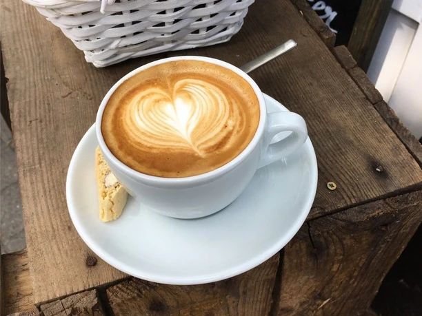 Eine Tasse Cappuccino mit Herzmuster im Schaum, serviert auf einem weißen Unterteller mit Keks.