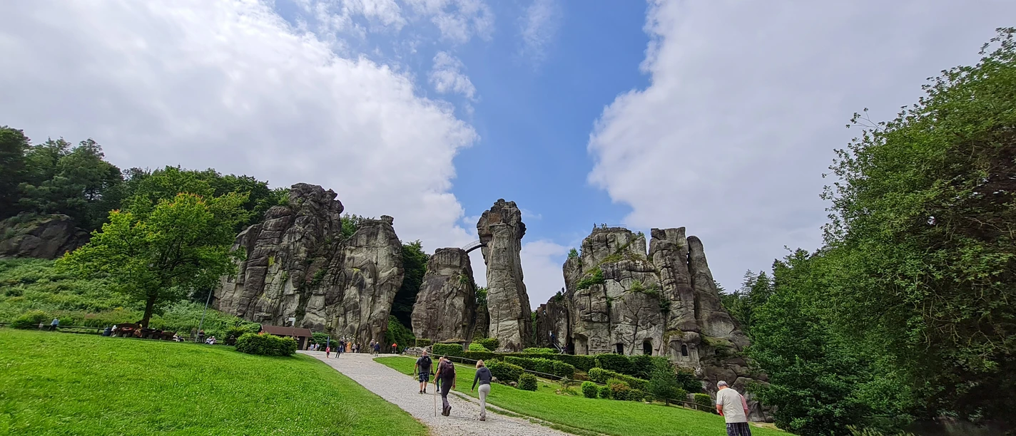 Externsteine_Front_®GesUndTourismus Horn-Bad Meinberg GmbH.jpg Gruppe von Menschen spaziert auf Kiesweg zu massiven Sandsteinfelsen, umgeben von grüner Natur.