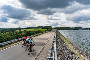 Staudamm Bever-Talsperre Drei Radfahrer fahren auf einem asphaltierten Weg entlang eines Stausees unter bewölktem Himmel.