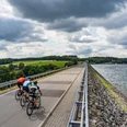Staudamm Bever-Talsperre Drei Radfahrer fahren auf einem asphaltierten Weg entlang eines Stausees unter bewölktem Himmel.