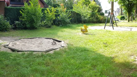 Grüne Parklandschaft mit Sandkasten, Enten-Wippspielzeug und Schaukel. Umgeben von Bäumen und Hecken.