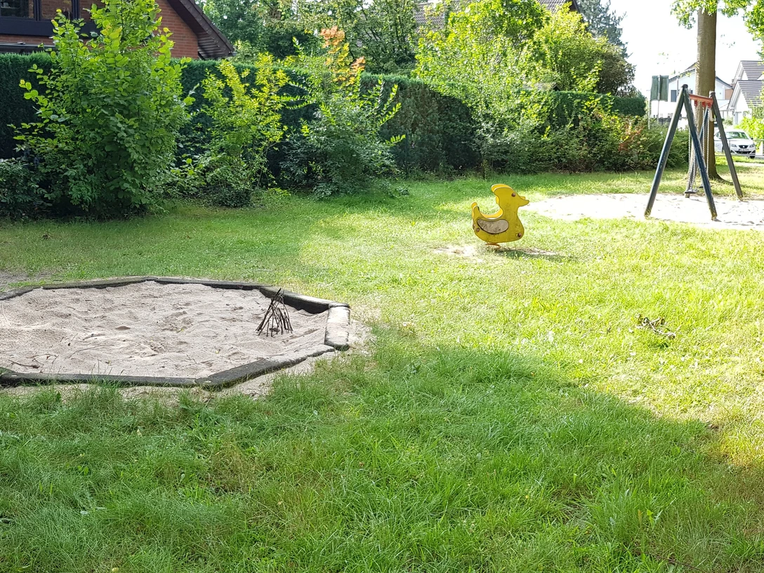 Grüne Parklandschaft mit Sandkasten, Enten-Wippspielzeug und Schaukel. Umgeben von Bäumen und Hecken.