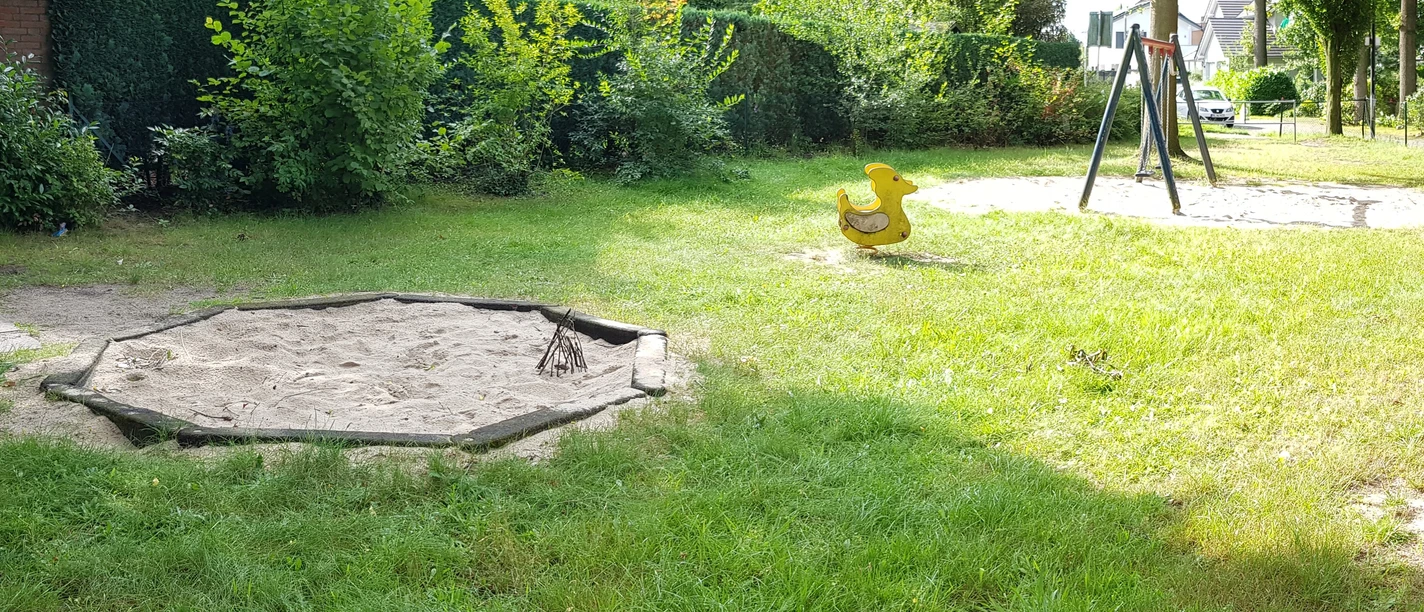 Spielplatz Finkenweg Grüne Parklandschaft mit Sandkasten, Enten-Wippspielzeug und Schaukel. Umgeben von Bäumen und Hecken.
