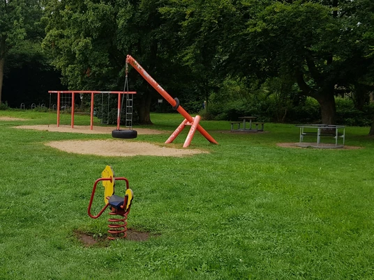 Spielplatz Grillenstraße groß Kinderspielplatz mit grüner Wiese, Schaukeln, Klettergerüst und Bäumen im Hintergrund.