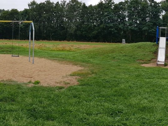 Spielplatz Grillenstraße groß Ein Spielplatz mit Schaukel und Rutsche, umgeben von Grasflächen und Bäumen im Hintergrund.